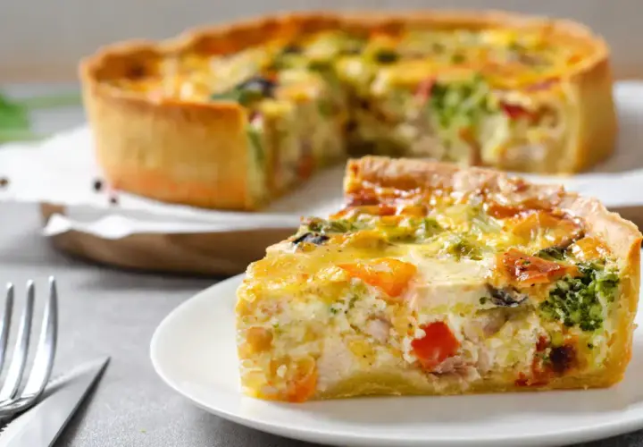 Une part de quiche savoureuse avec une pâte à tarte maison dorée, garnie de légumes et de poulet. Le reste de la tarte est flou en arrière-plan.