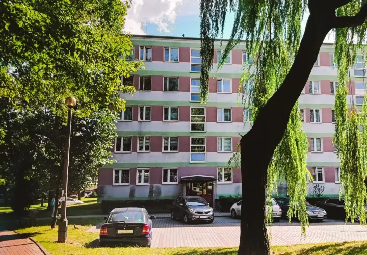 Blok mieszkalny z zielonymi i różowymi pasami, drzewa i zaparkowane samochody.
