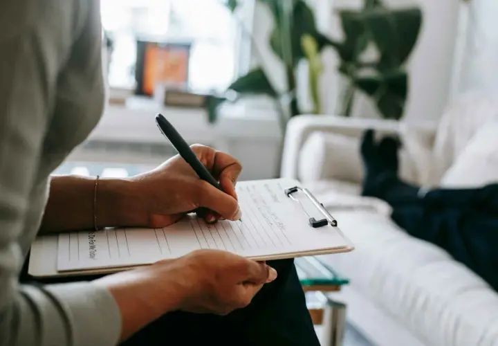 Mani che scrivono su un blocco note, mentre una persona è sdraiata su un divano. Un momento di riflessione per capire se per l'ansia meglio psicologo o psichiatra.