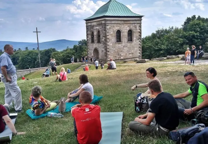 Ludzie odpoczywają na trawie pod kamienną wieżą i krzyżem. Trasa na Święty Krzyż przyciąga turystów.