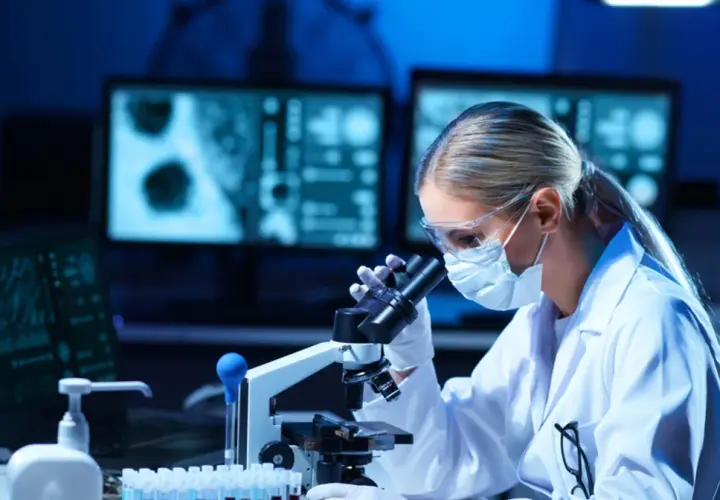 Diagnosta laboratoryjny w okularach i masce analizuje próbki pod mikroskopem. Na ekranach komputera widać wyniki badań.
