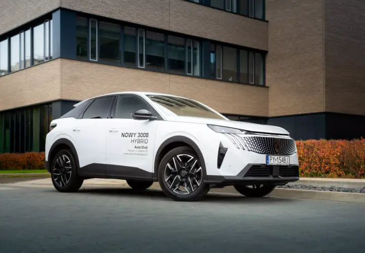 Biały samochód na e, nowy Peugeot 3008 Hybrid, zaparkowany przed nowoczesnym budynkiem z ceglaną elewacją.