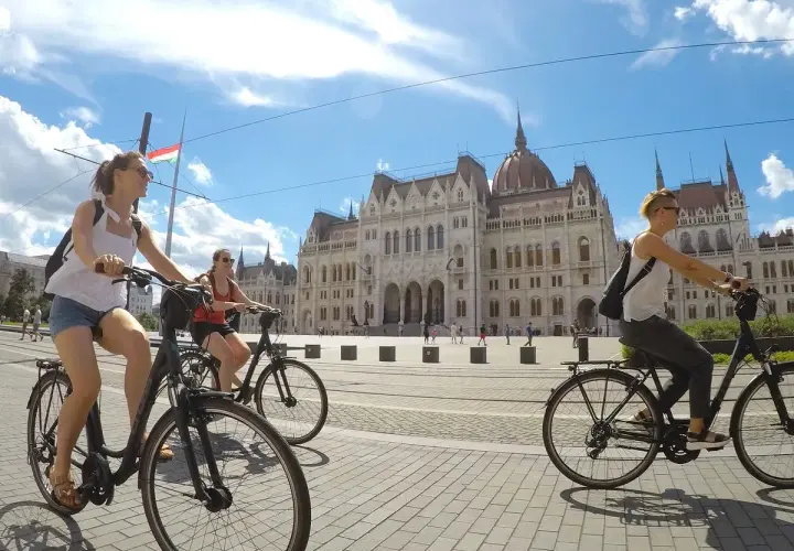 Friends enjoying Budapest bike tours, cycling past the Parliament building under a bright, cloudy sky.
