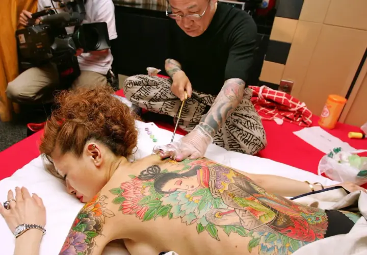 Mistrz tatuażu tworzy tradycyjne tatuaże w Japonii na plecach kobiety. W tle operator kamery dokumentuje proces.