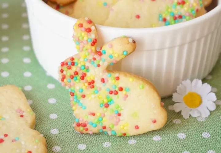 Bunte Osterkekse zum Ausstechen, verziert mit bunten Streuseln, liegen auf einem grünen Tupfentuch. Ein süßer Osterhase steht im Vordergrund.