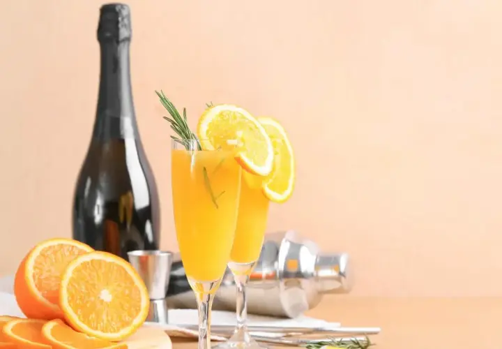 Dwa kieliszki z pomarańczowym drinkiem, ozdobione plasterkami pomarańczy i rozmarynem. Obok butelka, shaker i świeże pomarańcze. Wódka łza pszenicy idealnie pasuje do takiego orzeźwienia.
