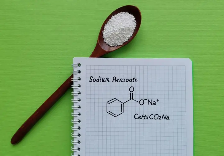 Drewniana łyżka z białym proszkiem, obok notatnik z napisem "Sodium Benzoate" i jego wzorem chemicznym.