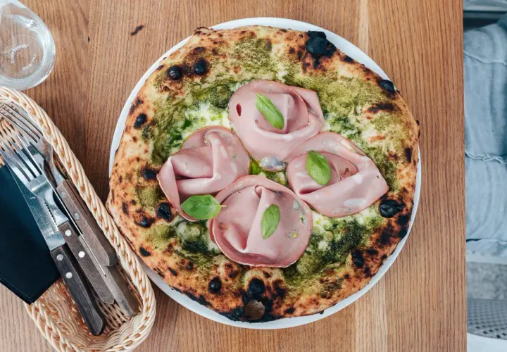 Pizza z mortadelą i pistacjami, zielonym pesto i listkami bazylii. Obok koszyk z widelcem i nożem.