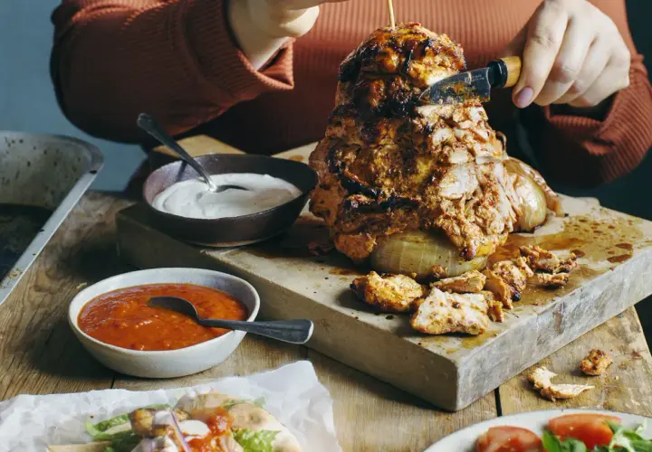 En person skär av en bit från en saftig kyckling shawarma som roterar på ett spett. Tillbehör som vitlökssås och en röd sås finns bredvid.