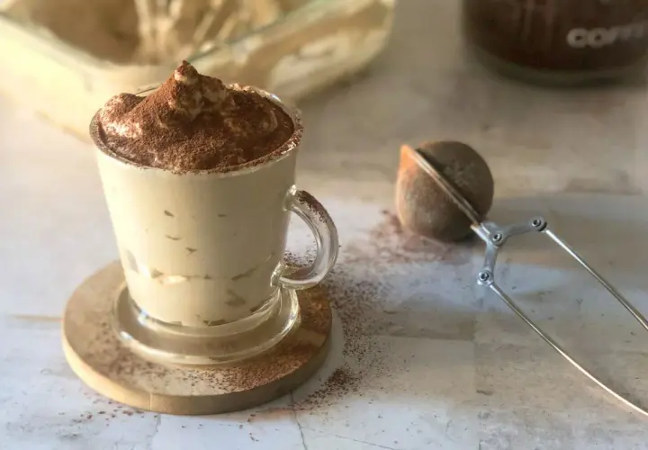 Deliziosa crema al caffè in una tazza di vetro, spolverata di cacao. Accanto, un colino per dolci e un barattolo di caffè.