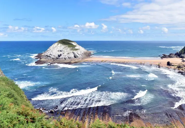 Olas rompen en las **playas limpias de Cantabria**, con un islote rocoso y vegetación verde.