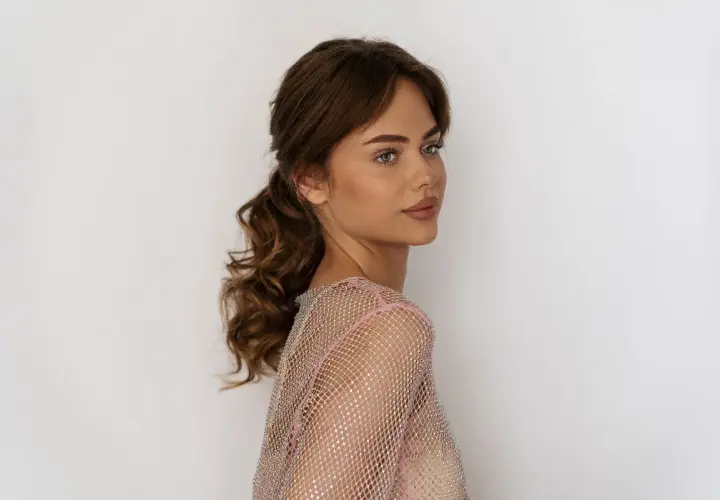 Semirecogidos peinados de madrina media melena: una mujer con cabello castaño ondulado recogido en una cola baja, luciendo un vestido brillante.
