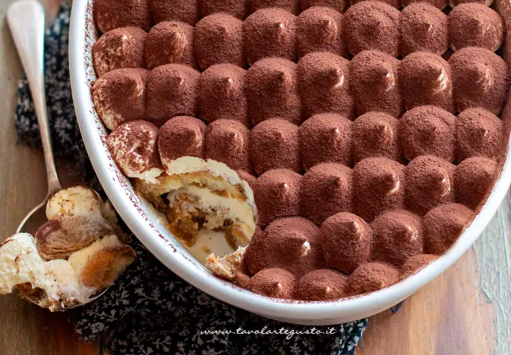 Un invitante tiramisù in una teglia bianca, cosparso di cacao. Una porzione è già stata servita su un cucchiaio, mostrando gli strati cremosi.