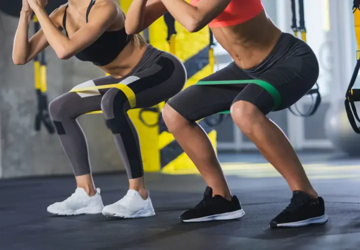 Dwie kobiety wykonują przysiady z gumami oporowymi na udach. Ćwiczenie to angażuje mięśnie podobnie jak maszyna hack squat, wzmacniając nogi i pośladki.