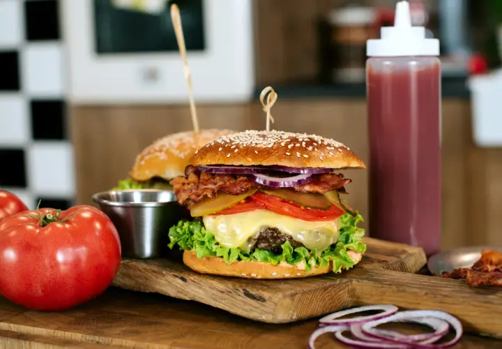 Soczysty burger wołowy z bekonem, serem i warzywami na desce, obok pomidorów i sosu.