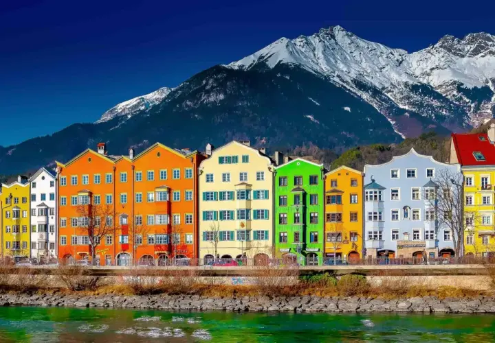 Colorful buildings line the Inn River, a vibrant scene on the journey from St. Moritz to Innsbruck, with snow-capped mountains in the background.