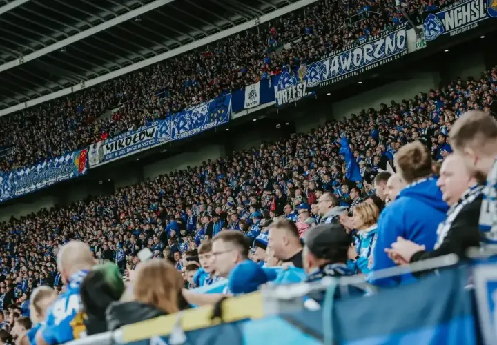 Tłum kibiców na stadionie, w niebieskich barwach, z transparentami "Jaworzno", "Mikołów", "Chorzów". Wszyscy w zgodzie dopingują swoją drużynę.