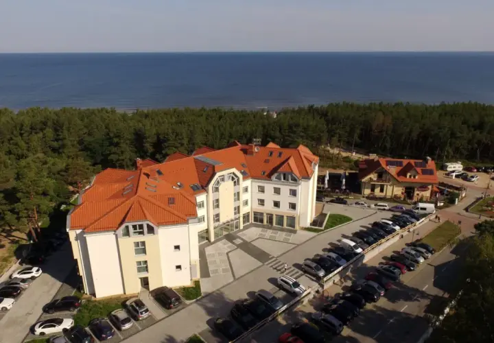 Hotel blisko morza, idealny na wypad z Śląska. Widać parking pełen aut, las i błękitną wodę.