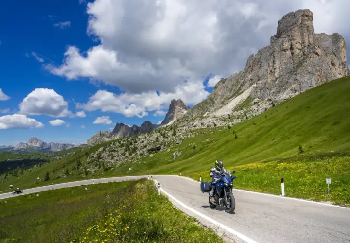 Motociclista percorre la strada cimina tra prati verdi e montagne imponenti sotto un cielo azzurro con nuvole bianche.