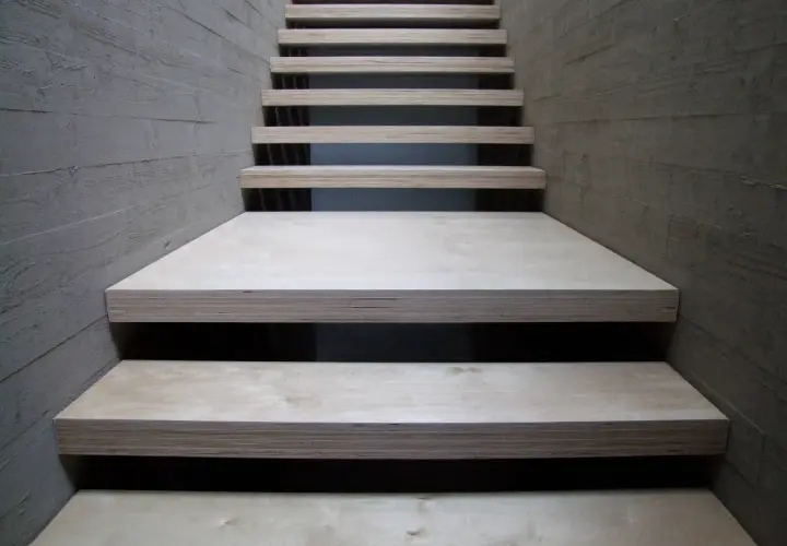 Una scala moderna con gradini in legno compensato, materiale resistente e versatile, fiancheggiata da pareti in cemento grezzo.