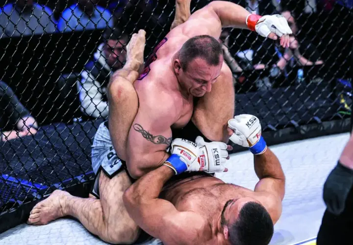 Intensywna walka Pudziana w klatce MMA. Jeden zawodnik klęczy, drugi próbuje go obalić.