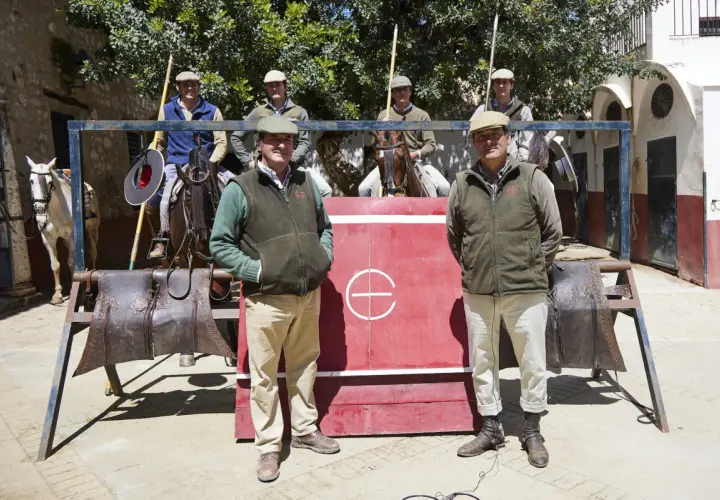 Hombres con gorras y chalecos posan frente a una barrera roja. Uno de ellos, Julio de la Puerta, sonríe. Caballos y equipo de equitación completan la escena.