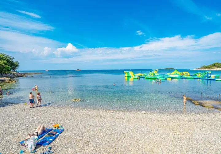 Słoneczna plaża Bijela Uvala z kamyczkami, turkusową wodą i dmuchanymi atrakcjami wodnymi. Ludzie odpoczywają i kąpią się.