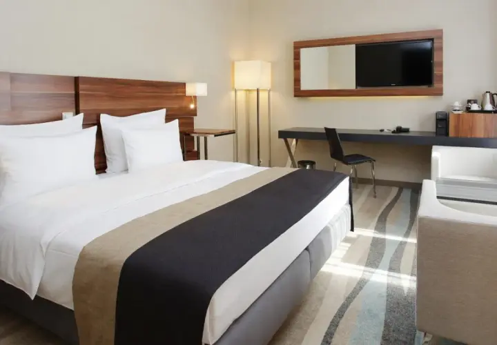 Nowoczesne wyposażenie pokoi hotelowych: wygodne łóżko, biurko z TV, fotele i stolik.