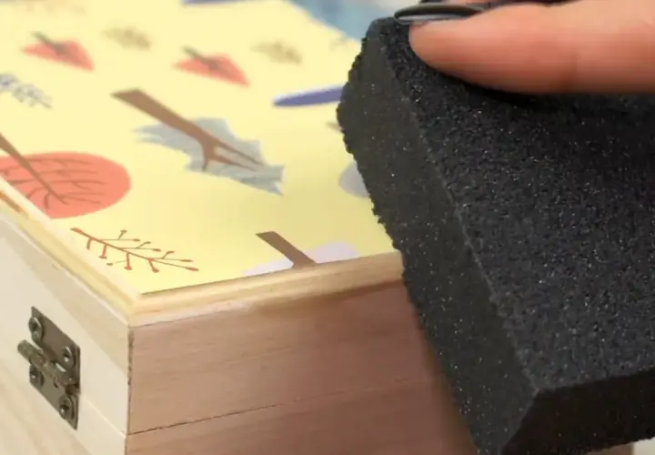 Aplicando pegamento para saber como pegar papel en madera, con una esponja sobre una caja decorada con hojas.