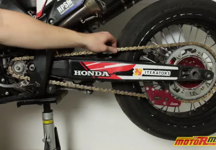 Ręka reguluje naciąg łańcucha w motocyklu Honda. Widać zębatkę, wahacz i tylne koło.