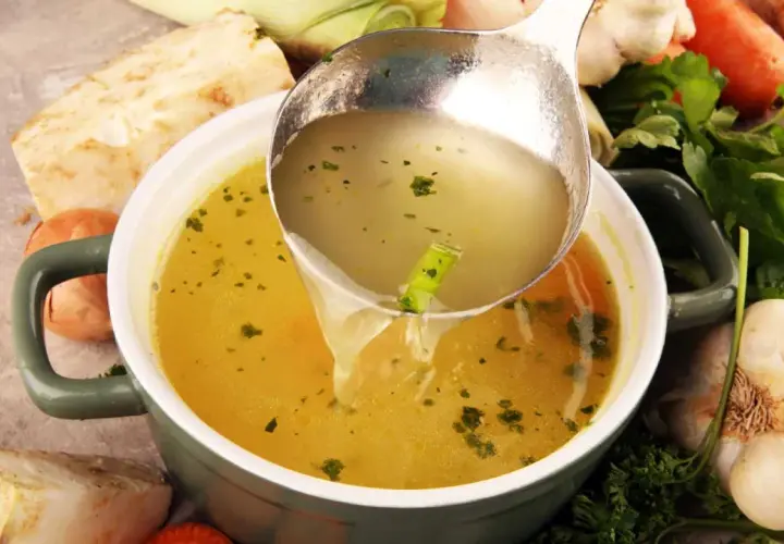 Un cucharón sirve un caldo depurativo, rodeado de verduras frescas como apio, zanahoria y ajo.