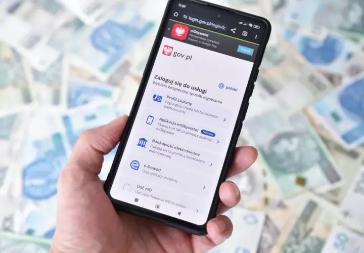 Dłoń trzyma smartfon z ekranem logowania do serwisu gov.pl. Dostępne opcje to m.in. Profil zaufany przez bankowość elektroniczną.