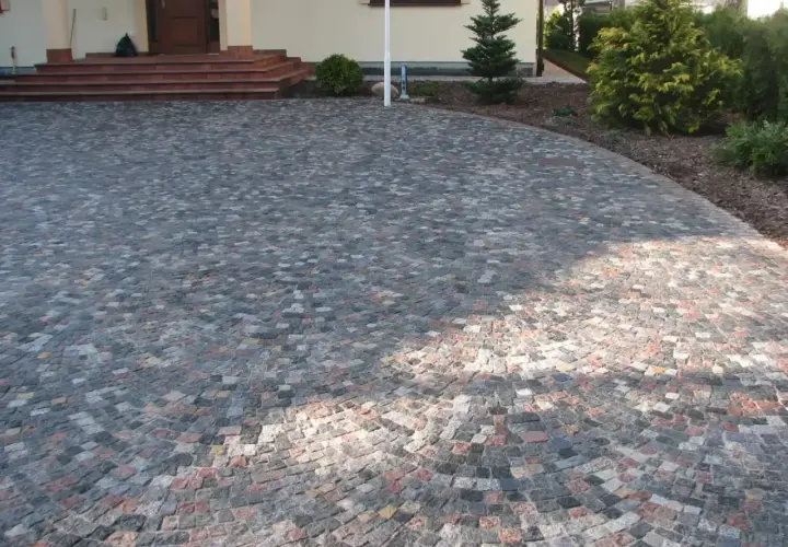 Piękny podjazd z kamienia, ułożony w łuk, prowadzi do drzwi domu. Wokół rosną drzewka i krzewy.