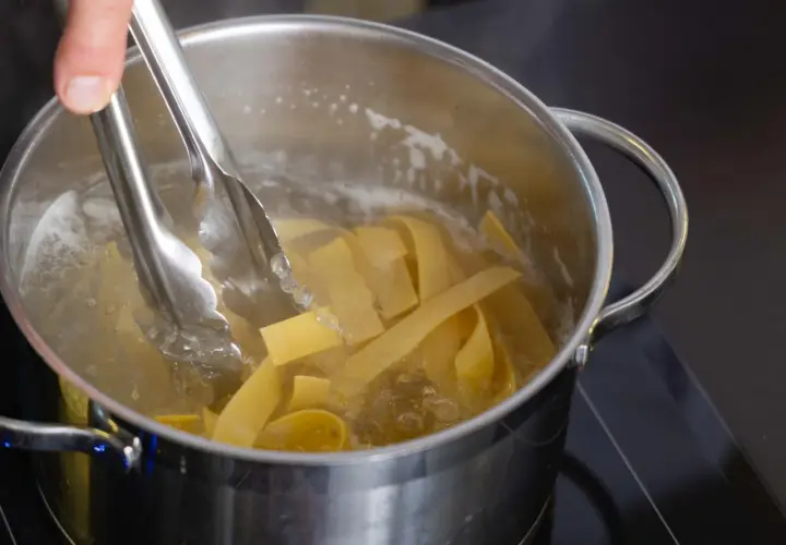 Szczypce wkładają szeroki makaron do wrzącej wody. Idealny moment, kiedy wrzucić makaron do wody, by był al dente.