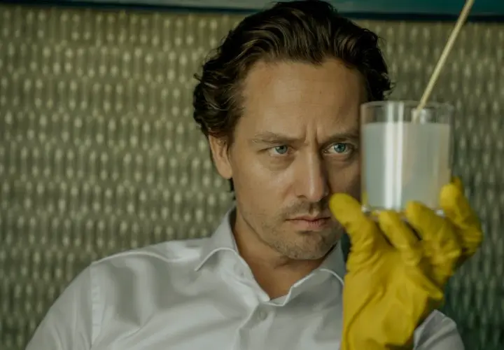 Ein Mann mit blauen Augen und gelben Handschuhen hält ein Glas mit weißer Flüssigkeit. Er ist Teil der "Achtsam Morden Reihe".