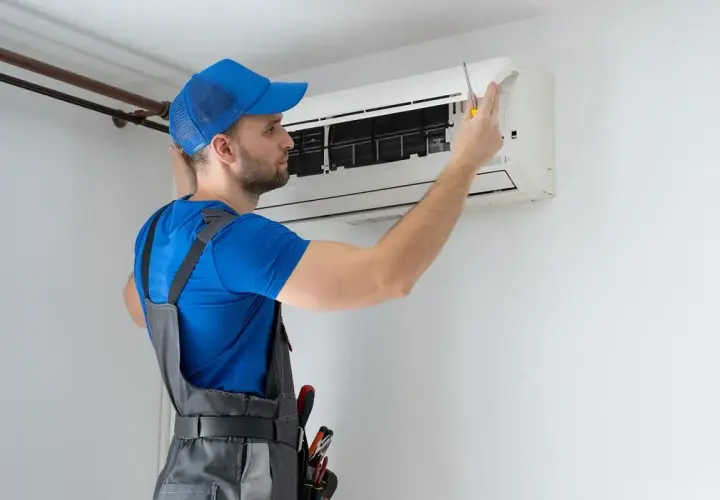 Technik w niebieskiej czapce i ogrodniczkach naprawia klimatyzator.