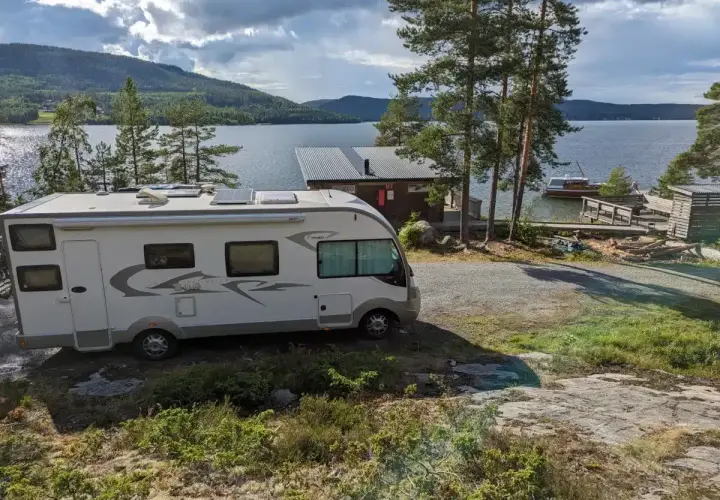 Un camping-car blanc est garé près d'un lac bordé de pins. Une petite cabane et un bateau sont visibles sur l'eau.