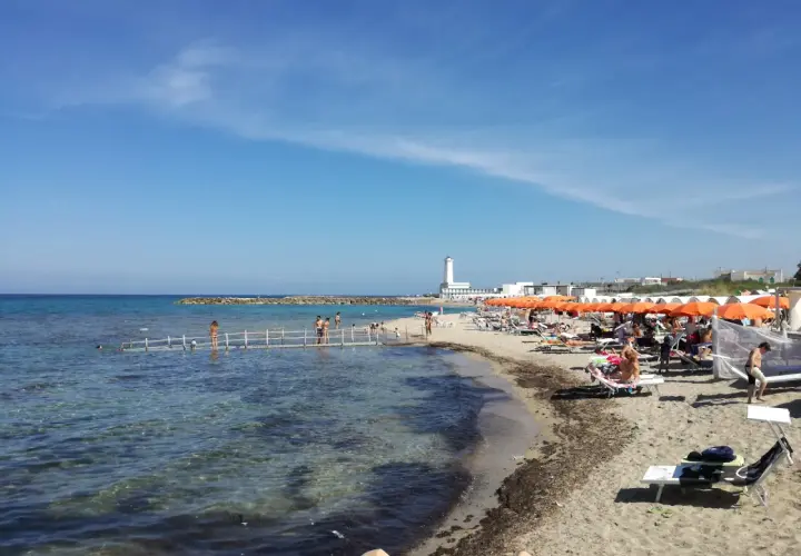 Plage animée à San Cataldo, Pouilles, avec des parasols orange, des baigneurs sur un ponton et un phare blanc en arrière-plan.