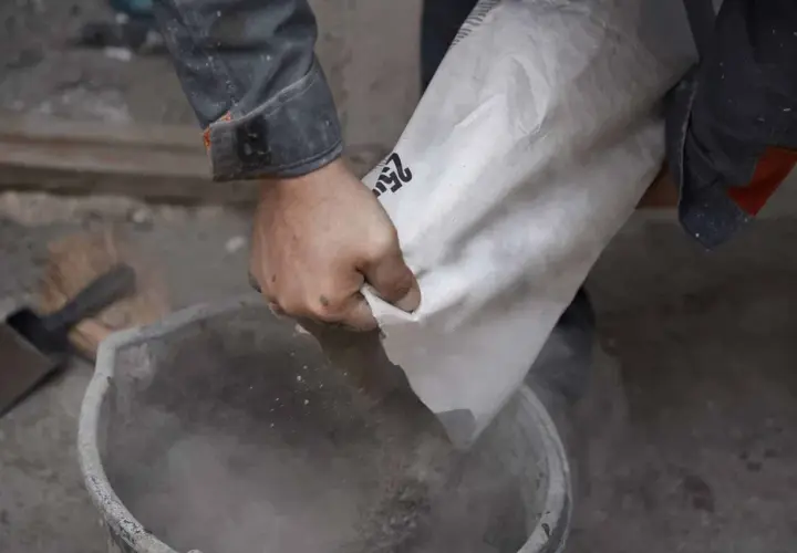 Ręce wsypują cement z worka do wiadra. Różne rodzaje cementu czekają na użycie.