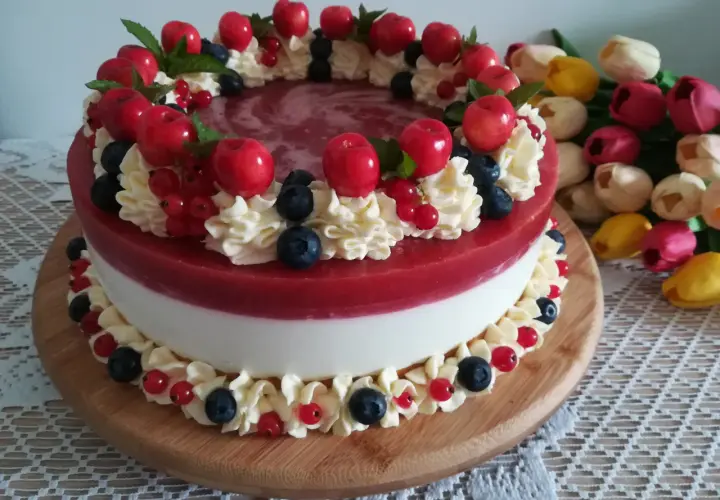 Wykwintny tort z warstwą białego kremu i błyszczącą, czerwona żelką truskawkową, ozdobiony bitą śmietaną, owocami i listkami mięty.