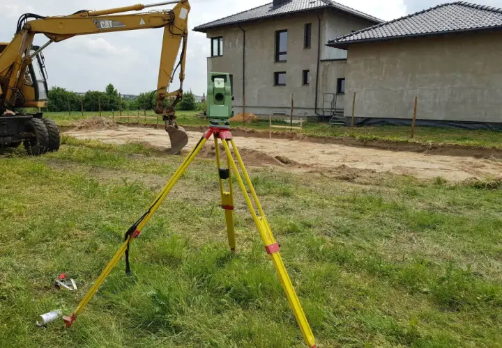 Geodeta z niwelatorem na statywie na placu budowy obok koparki i domów.