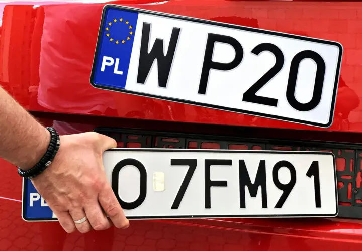 Ręka z bransoletką montuje tablicę rejestracyjną z napisem "O 7FM91" na czerwonym samochodzie. Powyżej widać inną tablicę "W P20".