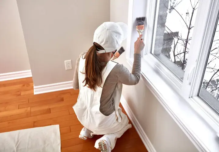 Donna con tuta da lavoro e cappellino bianco mentre è intenta a verniciare infissi in alluminio con un pennello.