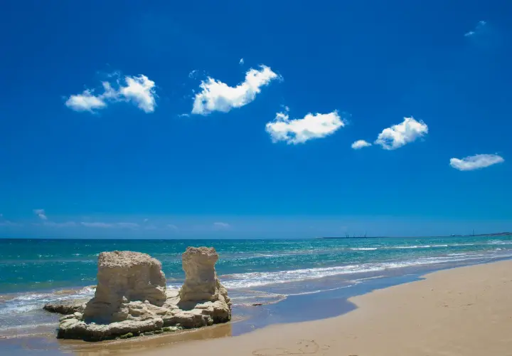 Spiagge libere Sicilia: rocce scolpite dal mare e sabbia dorata sotto un cielo azzurro con nuvole bianche.