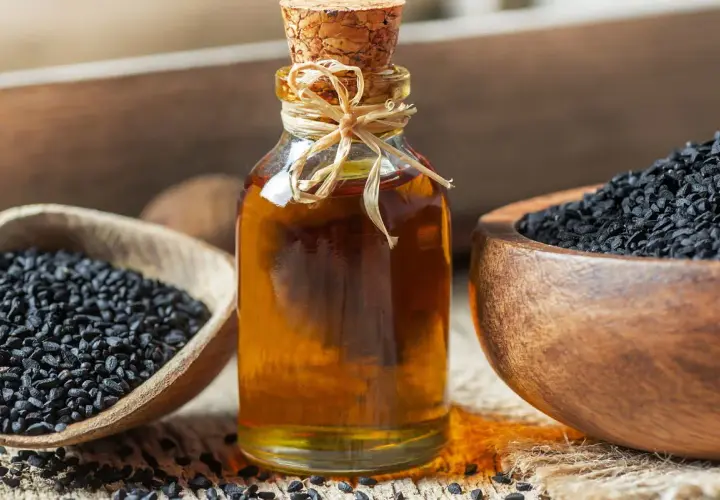 Naturalny olejek z czarnuszki na twarz w szklanej butelce, obok drewnianych miseczek z nasionami czarnuszki.