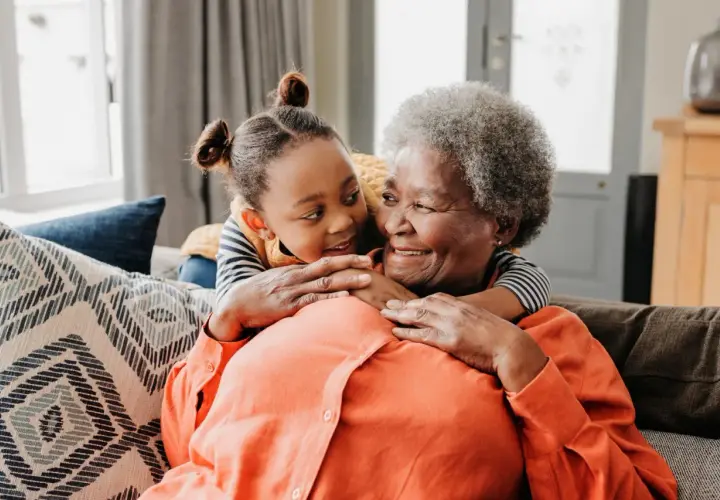 Ein süßer Moment zwischen Oma und Enkel. Das kleine Mädchen umarmt ihre Großmutter von hinten, beide lächeln sich liebevoll an.