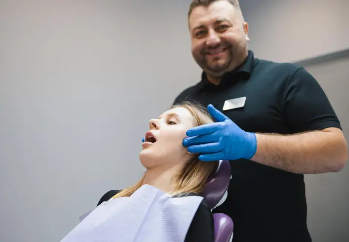 Uśmiechnięty dentysta w niebieskich rękawiczkach bada pacjentkę. Zespół Costena dba o zdrowie jamy ustnej.
