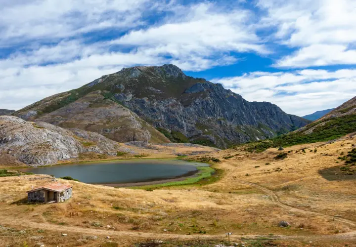 Pequeña cabaña de piedra junto a un lago ausente, rodeada de montañas escarpadas y prados secos bajo un cielo azul con nubes.