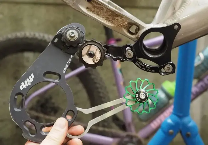 A hand holds a bike chain guide, part of a 1x drivetrain, near a bicycle frame. A green pulley wheel is also visible.