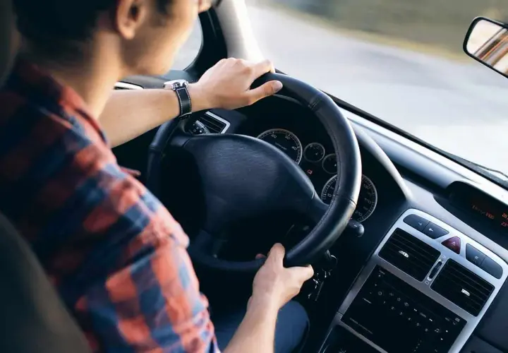 Uomo al volante di un'auto, con le mani sul volante. La sua camicia a quadri rossi e blu si abbina al cruscotto dell'eps auto.